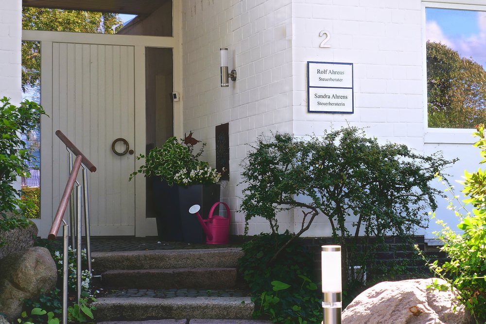 Eingang der Kanzlei in der Dreiangel 2 in Altenholz: weiß gestrichenes Haus mit heller Holztür und dem Schild der Steuerberatung